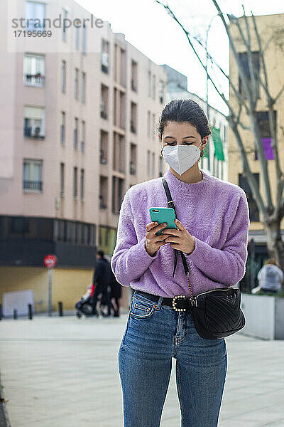 junge Frau mit KN95-Schutzmaske auf der Straße  die das Handy beobachtet