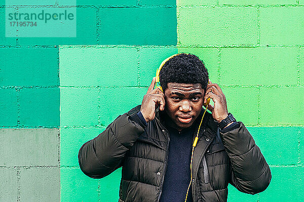 Schwarzer afroamerikanischer Junge auf grüner Hintergrundwand. Hören von Musik mit gelben Kopfhörern.