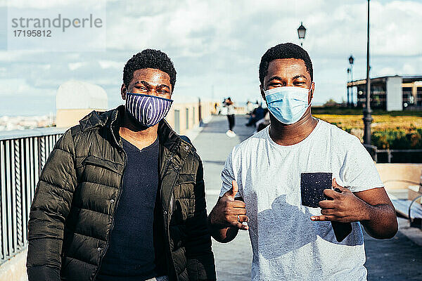 Porträt von zwei afroamerikanischen Freunden mit Gesichtsmaske  die fröhlich auf einem städtischen Platz posieren.