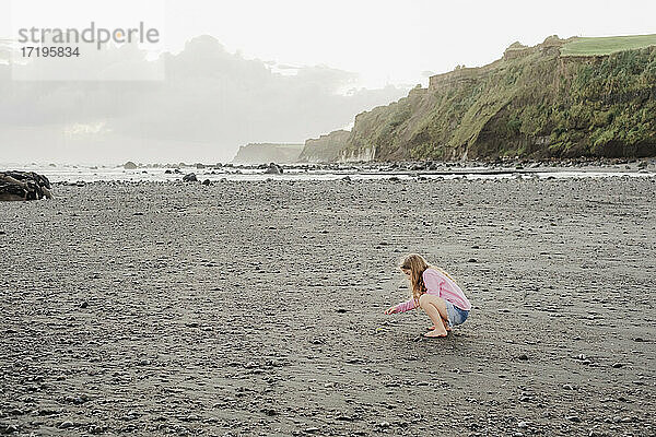 Junges Mädchen  das sich hinhockt und die Felsen am Strand betrachtet