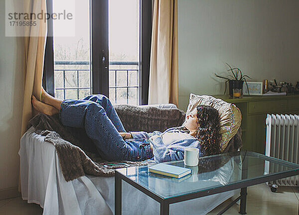 junge Frau mit lockigem Haar  die auf dem Sofa liegt und auf das Fenster schaut