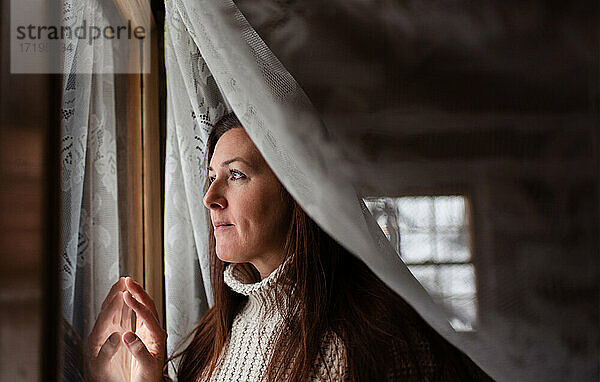 Attraktive Frau schaut durch ein Fenster hinter einem Spitzencuratin.