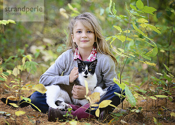 Junges blondes Mädchen sitzt im Wald mit schwarz-weißer Katze auf dem Schoß.