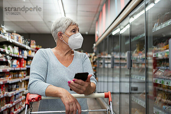 Kaukasische ältere Frau mit Gesichtsmaske und Telefon beim Einkaufen im Supermarkt