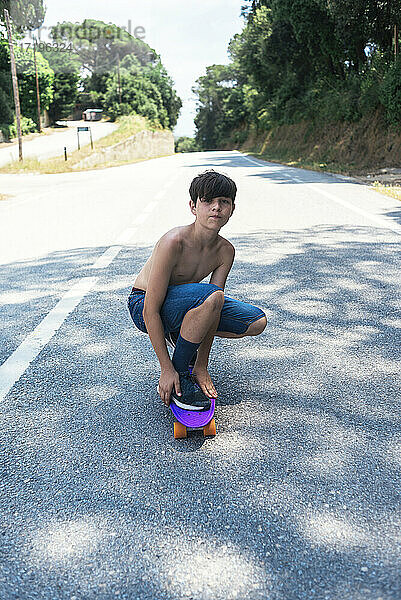 Porträt eines fröhlichen Teenagers ohne Hemd  der auf einer Bergstraße Skateboard fährt