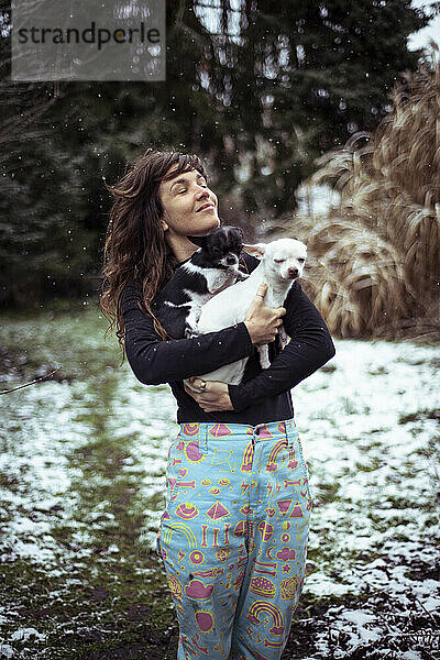 glückliche Frau steckt den Kopf in den Schnee und hält zwei kleine Hunde im Garten