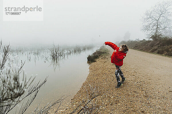 Junge in rotem Mantel wirft Stein in See gegen nebligen bedeckten Himmel