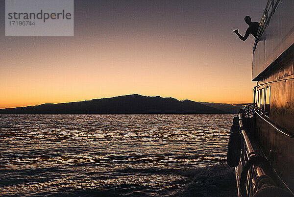 Die Silhouette eines Mannes zeigt einen Shaka auf einer Kreuzfahrt bei Sonnenuntergang mit Maui in der Ferne