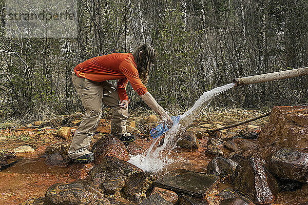 Seitenansicht eines Mannes  der eine Flasche mit Wasser füllt  das durch ein Rohr im Wald fließt