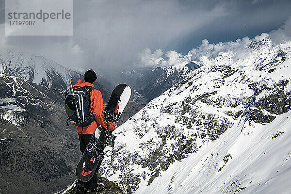 Mann mit Snowboard mit Blick auf Blick von schneebedeckten Berg gegen bewölkten Himmel