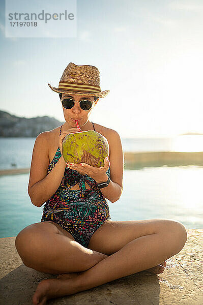 Glückliche Frau  die sich im Schwimmbad entspannt und Kokosnusswasser trinkt.