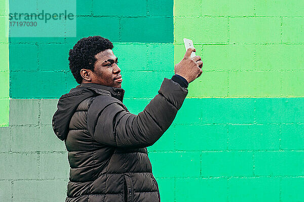 Junger schwarzer afroamerikanischer Junge  der ein Selfie mit seinem Mobiltelefon auf grünem Wandhintergrund macht.