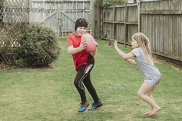 Junger Bruder und Schwester spielen mit Rugby-Ball im Hinterhof