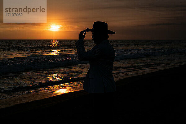 Silhouette eines Mannes mit Hut vor dem Meer bei Sonnenuntergang