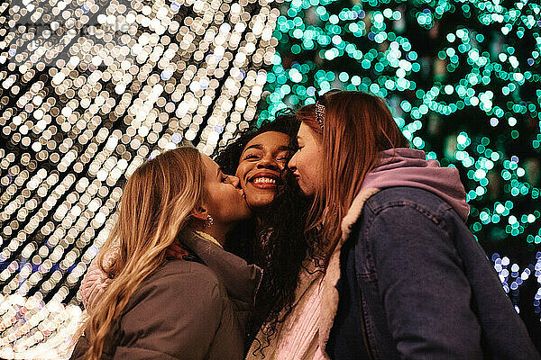 Freundinnen küssen junge Frau auf die Wangen bei Weihnachtsbeleuchtung