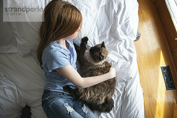 Junges glückliches rothaariges blauäugiges Mädchen auf weißem Bett mit Katze liegend.