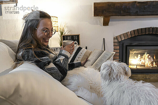 glückliche Frau zu Hause liegend auf Buch lesen und Kaffee trinken mit ihrem Hund