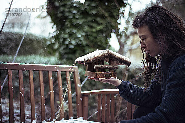 Asiatische Person im Schnee hält hölzernes Vogelhäuschen  um Samen in den Hinterhof zu geben