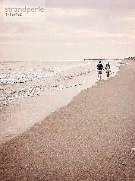 Mann und Frau gehen händchenhaltend an einem Strand in Richtung eines entfernten Kais