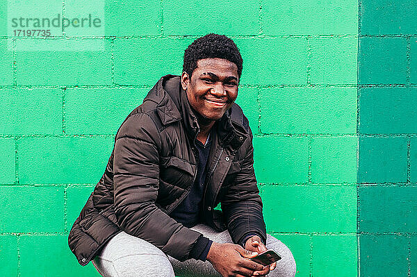 Junger schwarzer afroamerikanischer Junge hockt mit seinem Handy auf grünem Wandhintergrund.