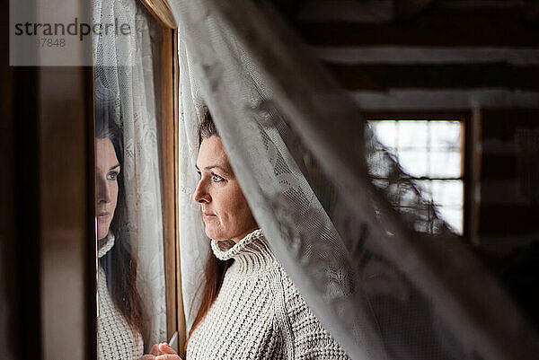 Attraktive Frau schaut durch ein Fenster hinter einem Spitzencuratin.