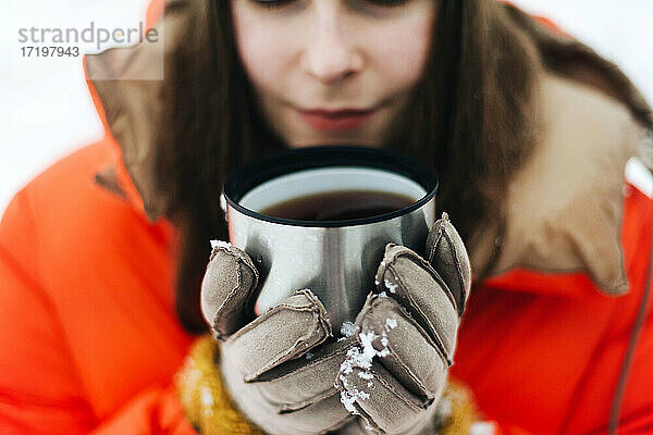Frau in orangefarbener Jacke trinkt heißen Tee im Freien im Winter.