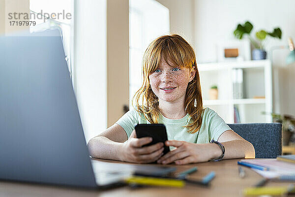 Lächelndes rothaariges Mädchen  das ein Smartphone hält und auf einen Laptop im Wohnzimmer schaut
