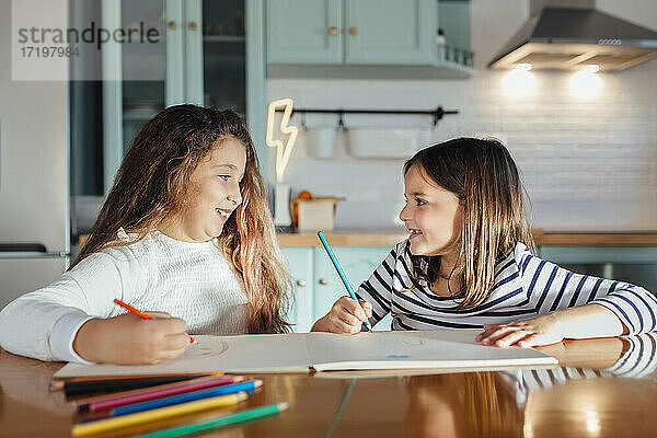 Lächelnde Mädchen schauen sich beim Malen mit Buntstiften auf Papier an  während sie am Esstisch in der Küche sitzen