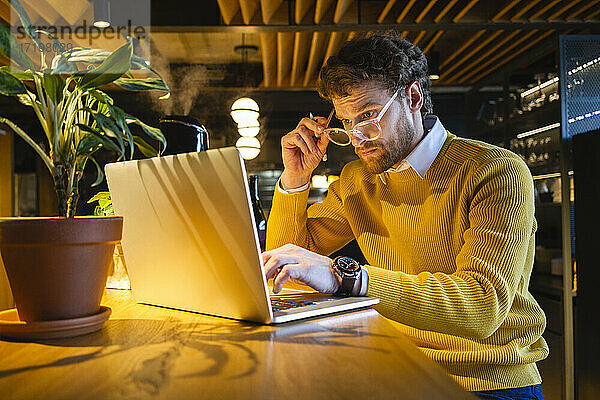 Männlicher Besitzer hält eine Brille in der Hand  während er in einem Café am Laptop arbeitet