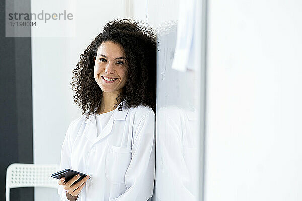 Lächelnde Ärztin mit Smartphone an der Wand lehnend in einer Klinik
