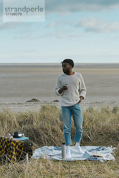 Junger Mann trinkt Tee  während er am Strand vor einem bewölkten Himmel steht