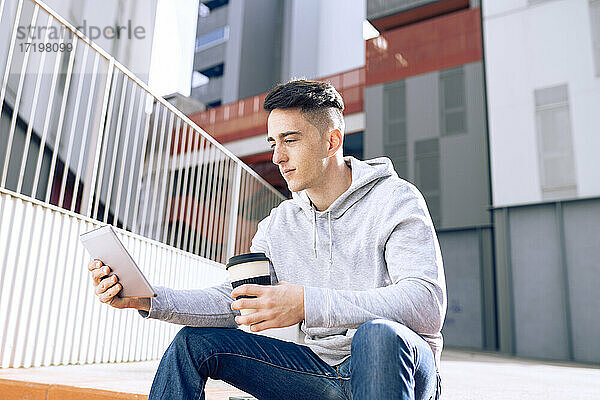 Junger Mann hält einen wiederverwendbaren Kaffeebecher  während er ein digitales Tablet vor einem Gebäude benutzt