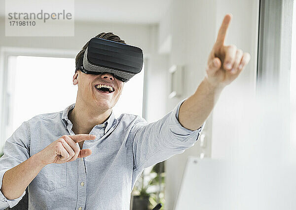 Fröhlicher männlicher Unternehmer gestikuliert  während er einen Virtual-Reality-Simulator im Heimbüro trägt