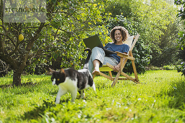 Glückliche Frau mit Laptop  die ihre Katze betrachtet  während sie auf einem Liegestuhl am Zitronenbaum sitzt