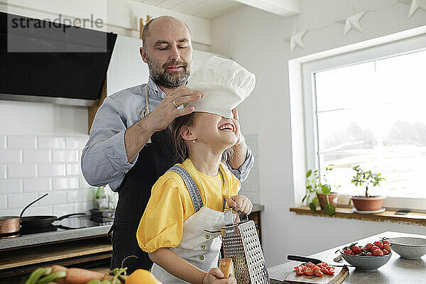 Vater setzt seiner Tochter die Kochmütze auf den Kopf  während er zu Hause in der Küche steht