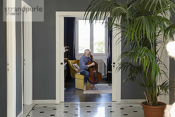 Älterer Mann spielt Cello  während er zu Hause auf einem Hocker sitzt