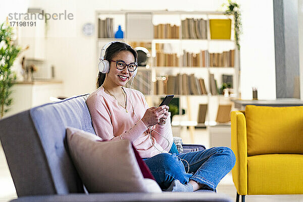 Junge Frau mit Mobiltelefon und Kopfhörern sitzt zu Hause auf dem Sofa