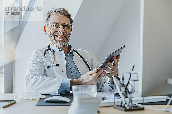 Männlicher Arzt  der ein digitales Tablet benutzt  während er in der Arztpraxis sitzt