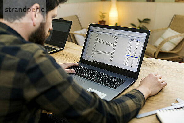 Männlicher Schreiner  der am Schreibtisch sitzend eine Zeichnung auf einem Laptop anfertigt