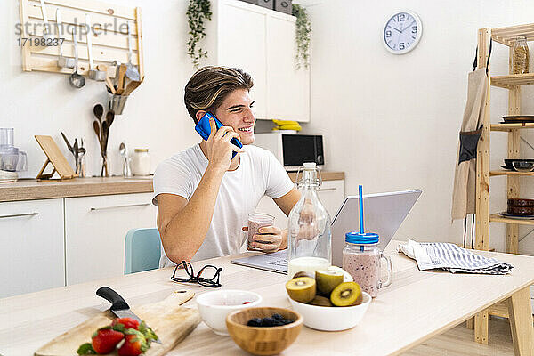 Lächelnder junger Mann  der mit dem Handy telefoniert  während er mit dem Laptop in der Küche zu Hause sitzt