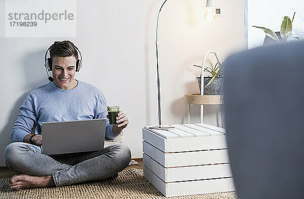 Lächelnder junger Mann mit drahtlosen Kopfhörern  der ein Video auf einem Laptop anschaut  während er im Wohnzimmer an der Wand sitzt
