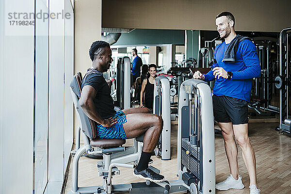 Lächelnde männliche Freunde unterhalten sich an einem Fitnessgerät im Fitnessstudio