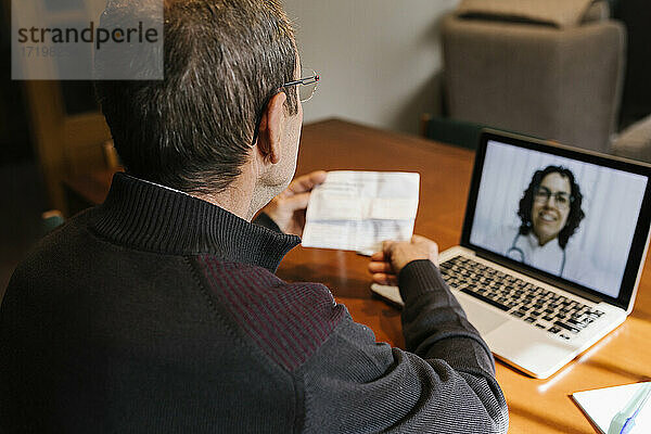 Ein älterer Mann hält ein Rezept in der Hand  während er per Videoanruf eine Ärztin zu Hause konsultiert  während COVID-19