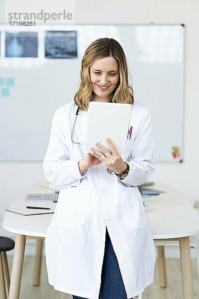 Lächelnder medizinischer Angestellter  der ein digitales Tablet benutzt  während er am Schreibtisch steht