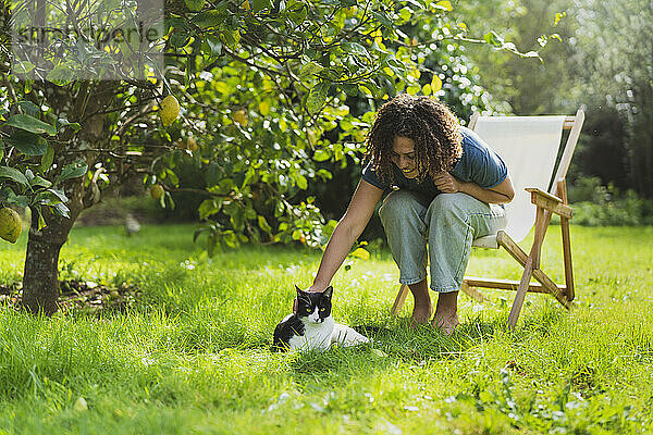 Lockenköpfige Frau streichelt Katze  während sie auf einem Stuhl im Garten sitzt