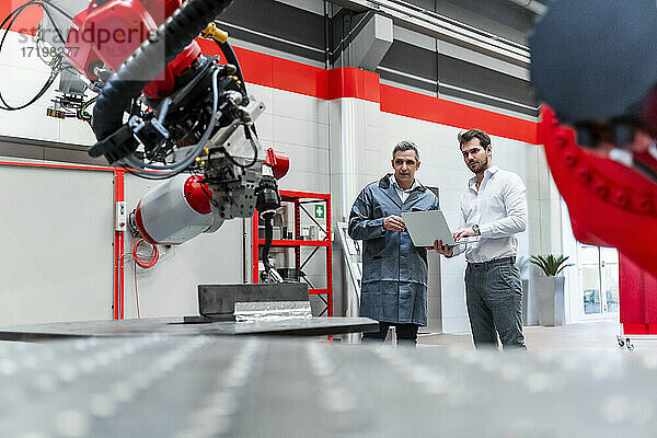 Männliche Ingenieure mit Laptop  die in einer Fabrik stehen und sich mit Robotik beschäftigen