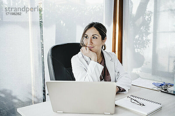 Lächelnde weibliche medizinische Fachkraft mit Hand am Kinn  die am Schreibtisch sitzt und träumt