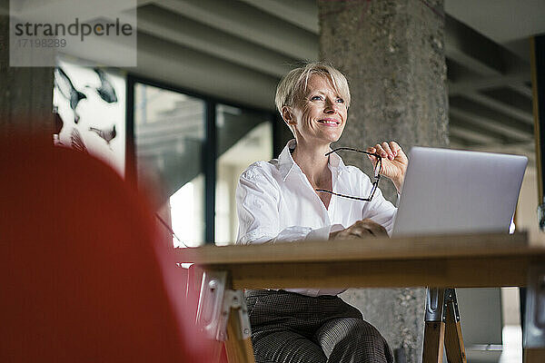 Lächelnde Geschäftsfrau mit Laptop  die eine Brille hält  während sie am Schreibtisch im Heimbüro sitzt