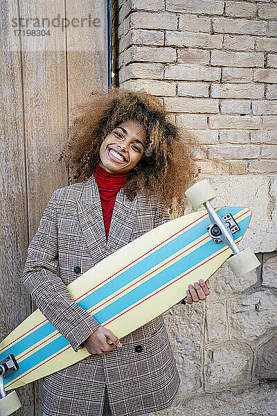 Lächelnde Afro-Frau mit Skateboard an der Wand stehend