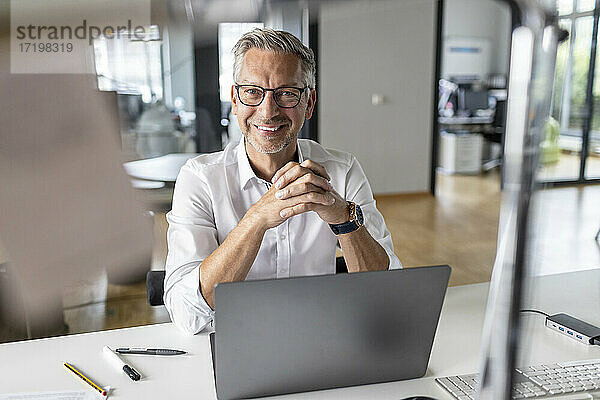 Lächelnder männlicher Unternehmer mit Laptop  der in einem Großraumbüro am Schreibtisch sitzt